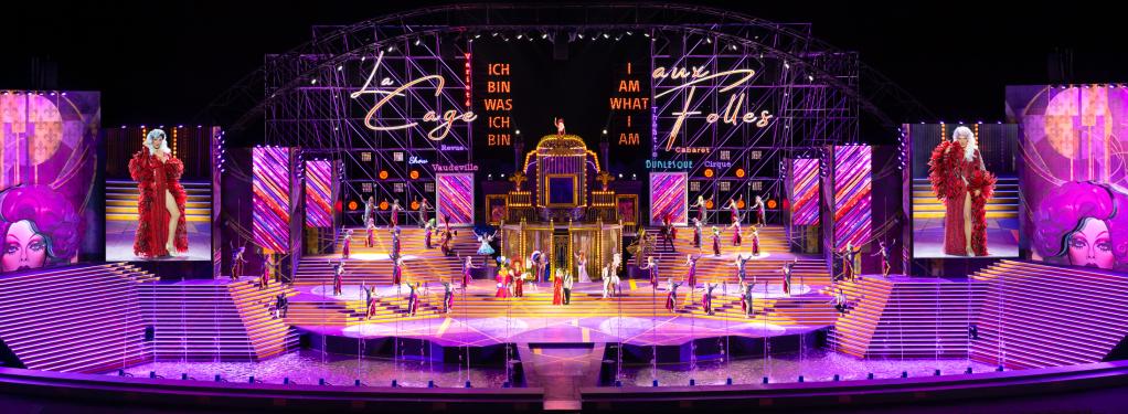 A wide stage with many steps. Pink and purple lighting with a La Cage Aux Folles neon sign in the background.